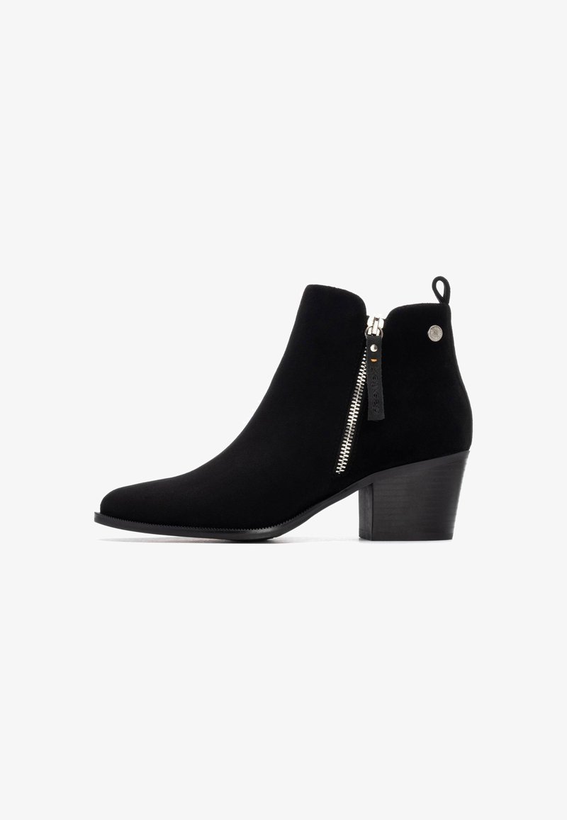 Botín de ante negro hasta el tobillo con punta afilada, detalle de cremallera lateral y tacón apilado. Presenta una pequeña lengüeta en la parte posterior.