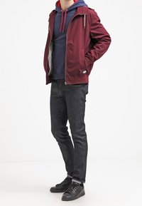 Giacca leggera borgogna con cappuccio, chiusura con zip e rivestimento grigio, indossata sopra una felpa blu navy con lacci rossi, abbinata a jeans scuri e sneakers nere.