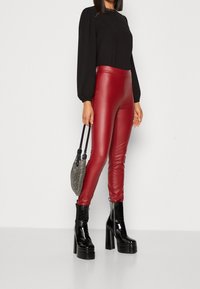 Femme portant un haut noir à manches longues, un pantalon en cuir rouge moulant, des bottines noires à plateforme, et tenant un petit sac à main argenté.