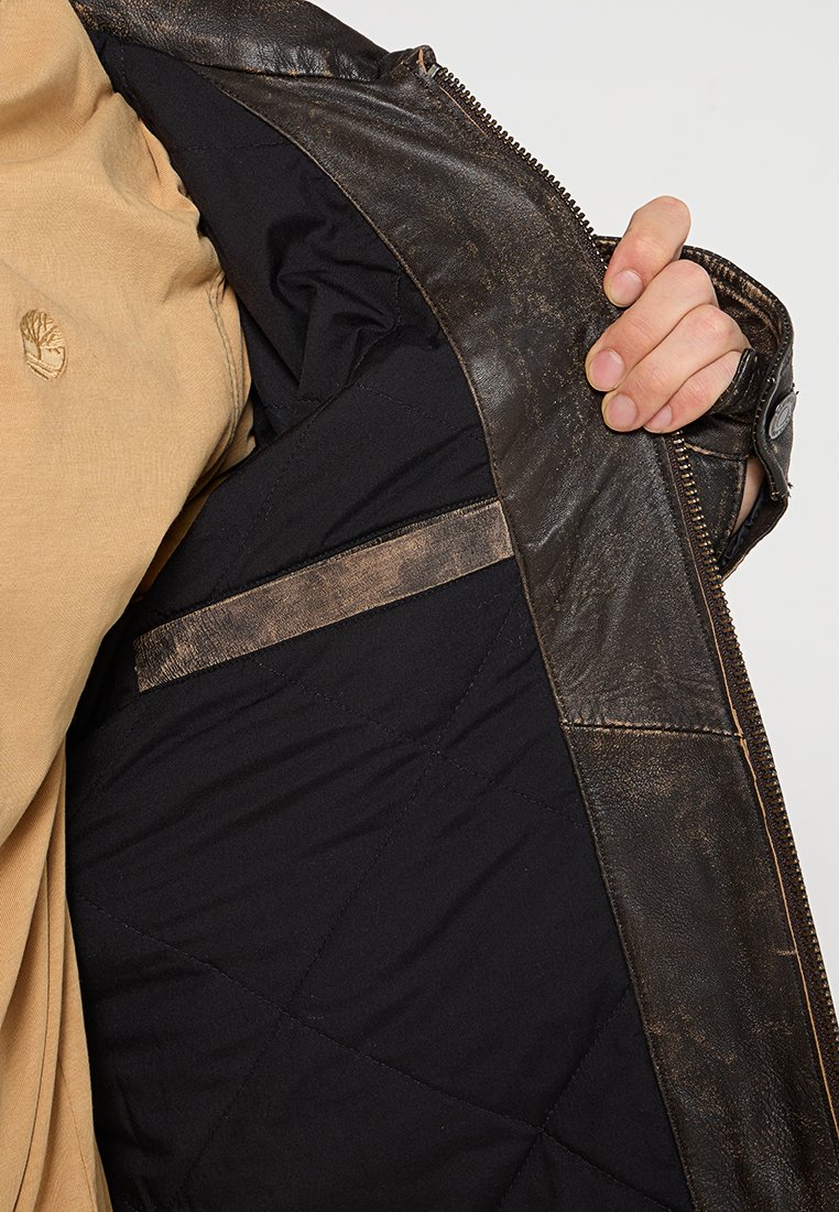 Brązowa skórzana kurtka z czarną pikowaną podszewką, z poziomą kieszenią i kontrastującymi detalami ściegów wzdłuż brzegów.