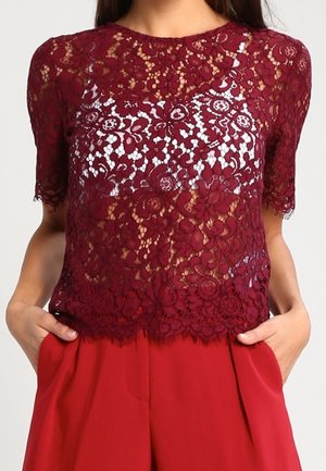 Femme portant un haut en dentelle florale bordeaux à manches courtes par-dessus une couche blanche, associé à un pantalon rouge taille haute, mains dans les poches.