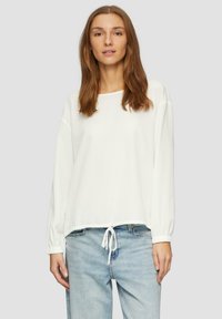 Haut blanc à manches longues en tissu texturé, avec des détails côtelés et une taille à cordon. Associé à un jean bleu clair.
