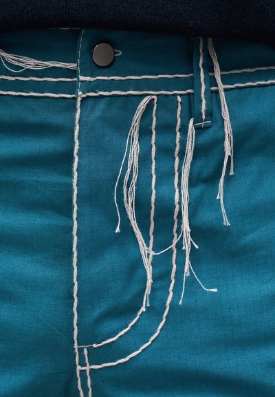 Τυρκουάζ ύφασμα με λευκές ραφές που μοιάζουν με τσέπη, μερικές χαλαρές λευκές κλωστές και ένα σκούρο κουμπί στο πάνω άκρο.