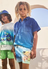 Due ragazzi sono in piedi di fronte a un muro bianco. Uno indossa una polo a degradé blu e pantaloni corti con un motivo chiaro; l'altro ha una t-shirt con una grafica e pantaloni corti verdi.
