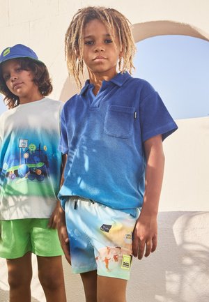 Due ragazzi sono in piedi di fronte a un muro bianco. Uno indossa una polo a degradé blu e pantaloni corti con un motivo chiaro; l'altro ha una t-shirt con una grafica e pantaloni corti verdi.