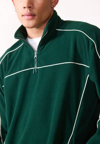 Grøn fleece pullover med kvart lynlås og hvid piping. Har en høj krave, lynlås lukning og blødt, struktureret stof.