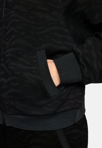 Schwarzer Sweatshirt mit Reißverschluss und strukturiertem Muster, ausgestattet mit einer Seitentasche sowie gerippten Bündchen und Saum. Das Material hat ein weiches, leicht glänzendes Finish.