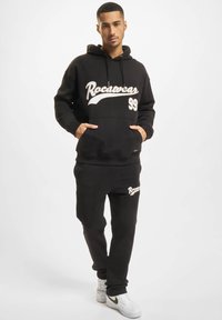 Melna kapuce un sporta bikses komplekts ar "Rocawear" logo baltā rakstā, ar priekšējo kabatu, kapuci ar aukliņām un ribotiem piedurknēm.