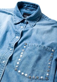 Chemise en denim bleu clair avec un col pointu, des boutons-pression sur le devant, et une poche soulignée par des clous métalliques sur la poitrine.