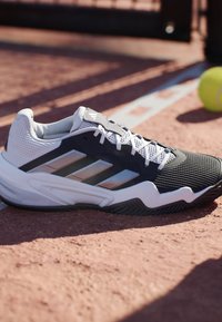 Sort og hvid sportssko med sølvstriber på en grus tennisbane nær en hvid linje og en gul tennisbold.