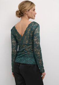 Top in pizzo teal a maniche lunghe con profondo scollo a V sul retro, decorato con motivi floreali e dalla vestibilità aderente. Abbinato a pantaloni in satin neri lisci.