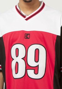 Camiseta roja, negra y blanca con el gran "89" en el frente, cuello en V con acentos a rayas y logo en el pecho. Fabricada con tejido texturizado.