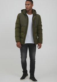 Pufjakke i mørkegrøn med et tekstureret olivengrønt panel, lynlås foran og hætte. Båret over en hvid skjorte og parret med mørke jeans og sneakers.