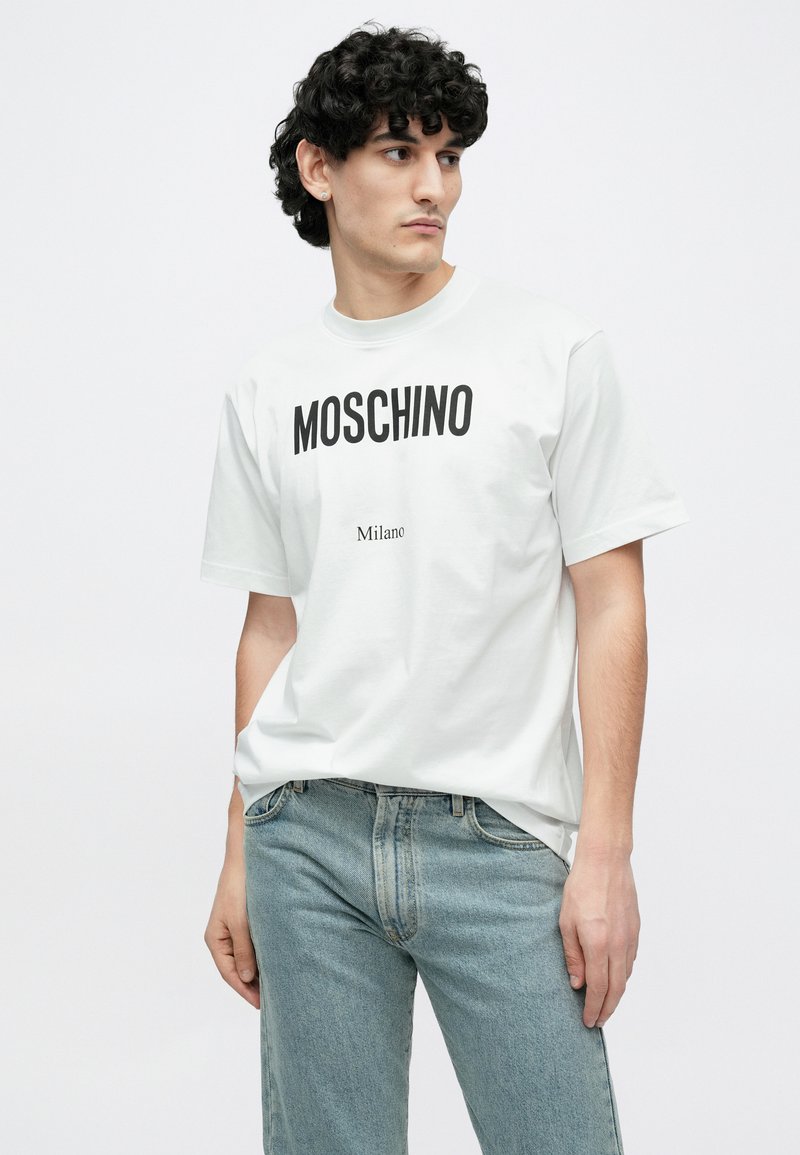 Hvid t-shirt med korte ærmer med det markante logo "MOSCHINO" i sort på brystet, med "Milano" trykt nedenunder i mindre tekst.