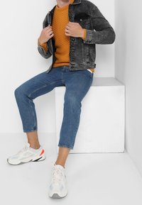 Personne portant un pull moutarde, une veste en jean noire, un jean court bleu et des baskets blanches assise sur un cube blanc dans une pièce blanche.