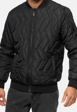Homme portant une veste bomber matelassée noire avec une fermeture éclair à l'avant, des poignets et une ceinture côtelés, et des poches latérales.