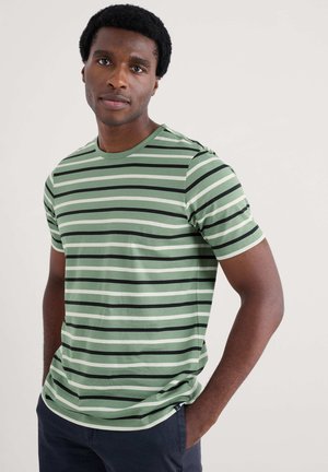 Junger Mann mit kurzen lockigen Haaren, der ein grün, schwarz und weiß gestreiftes T-Shirt und dunkle Hosen trägt, mit den Händen in den Taschen.
