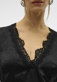 Sorte stoftop med en dyb V-hals og indviklet sort blonderafslutning. Glat tekstur med en subtil glans. Dekorativ tatovering synlig.