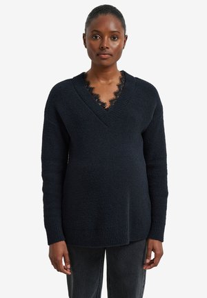 Frau trägt einen schwarzen Pullover mit V-Ausschnitt über einem Spitzenbesetzten Oberteil und dunkle Jeans, stehend vor einem schlichten weißen Hintergrund.