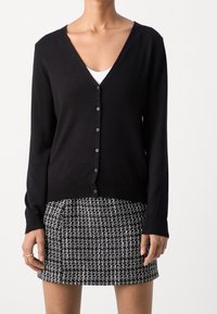 Svart kofta med V-ringning, långa ärmar och knappstängning, kombinerad med en kjol med svart-vitt mönster. Slät tygstruktur.