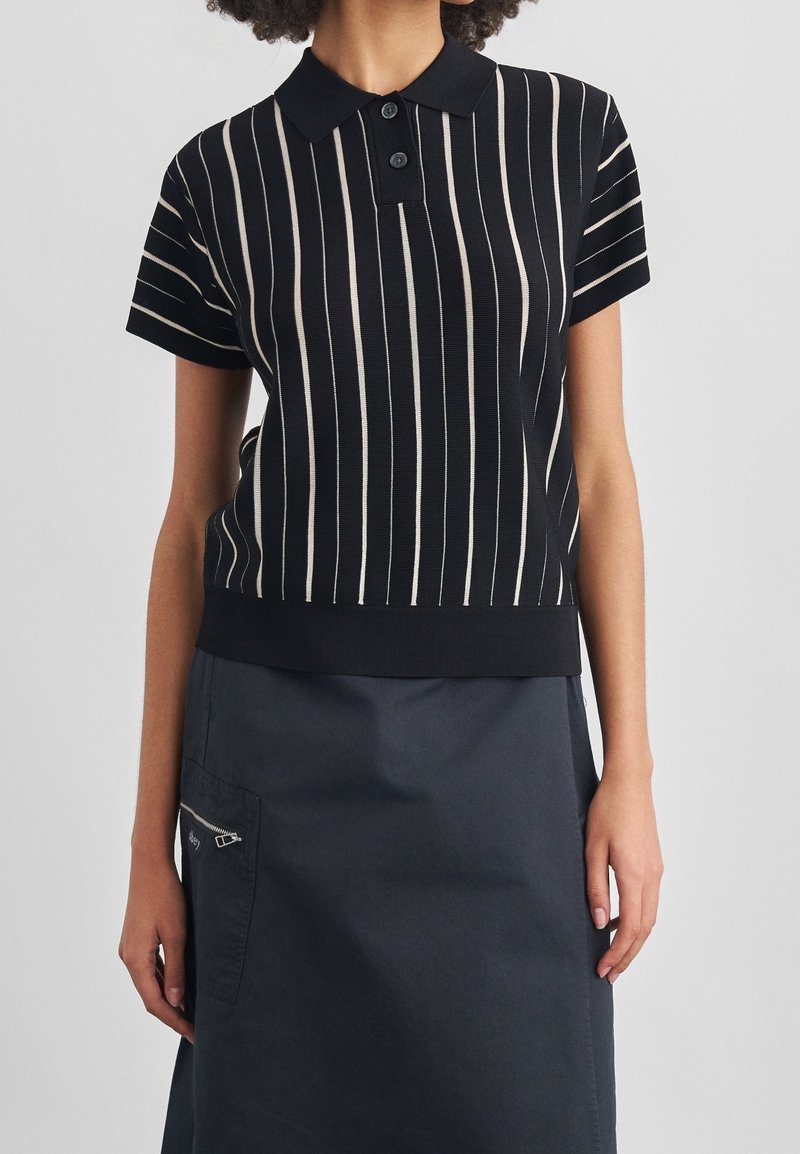 Femme portant un polo noir à manches courtes avec des rayures verticales blanches et une jupe sombre avec une poche latérale zippée.