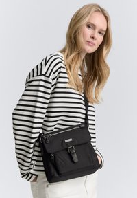 Schwarze Umhängetasche aus strapazierfähigem Stoff, mit Klappenverschluss, verstellbarem Schultergurt und silbernen Hardware-Details. Gestreifter Pullover-Hintergrund.