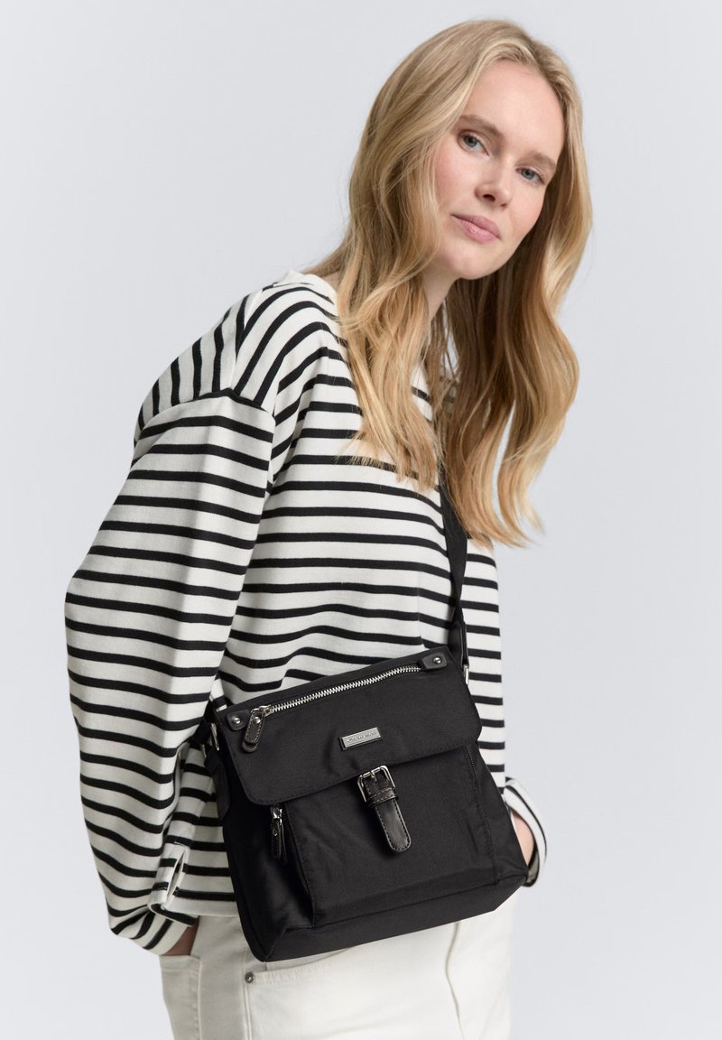 Borsa a tracolla nera realizzata in tessuto resistente, con chiusura a pattina, tracolla regolabile e accessori in metallo argentato. Sfondo a righe di un maglione.