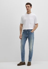 Weißes Kurzarm-T-Shirt mit Brusttasche, kombiniert mit hellblauen Jeans und braunen Wildleder-Loafers. Legerer Schnitt und klares Design.