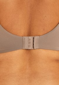 Rintaliivi vaaleassa beessissä, jossa on sileä kangastekstuuri, säädettävä hakaskiinnitys selässä ja siisti, minimalistinen muotoilu.