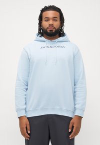 Man with beard and dreadlocks wearing light blue Jack & Jones hoodie and dark pants standing against a plain white background.