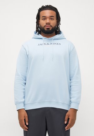 Man met een baard en dreadlocks, draagt een lichtblauwe Jack & Jones hoodie en donkere broek, staand tegen een vlakke witte achtergrond.