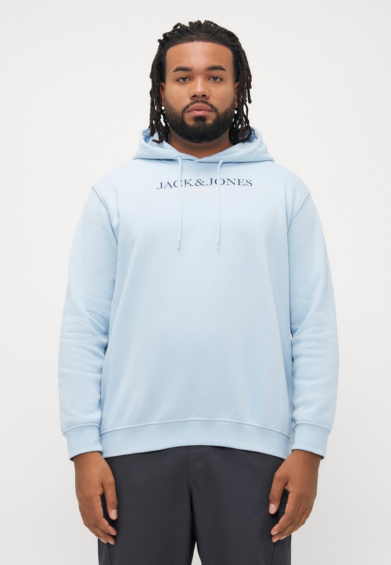 Man with beard and dreadlocks wearing light blue Jack & Jones hoodie and dark pants standing against a plain white background.