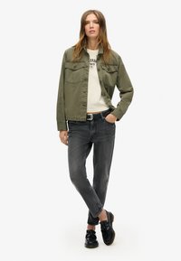 Olivno zeleni bombažni jopič z dvema žepoma na prsih, v kombinaciji z kremasto srajco in sivojeans hlačami. Model nosi črne mokasinke in stoji samozavestno.