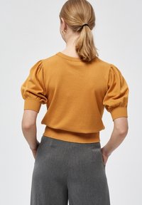Žena s blond copem, oblečená v hořčicovém svetr s krátkým rukávem a nadýchanými rukávy, a šedých kalhotách s vysokým pasem, s rukama spočívajícími na bocích.