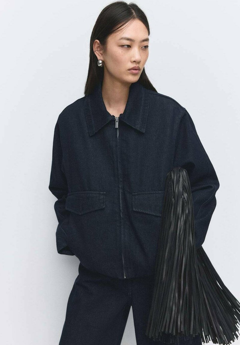 Dunkelblaue Jeansjacke mit einem kragenähnlichen Ausschnitt, Reißverschluss vorne und zwei großen Taschen. Dazu ein schwarzes Accessoire mit Fransen.