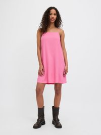 Rosa ermeløs kjole med firkantet hals, laget av myk stoff. Modellen har på seg svarte ankelsko, som fremhever det avslappede utseendet.
