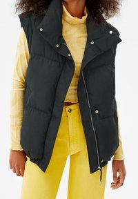 Black puffer vest with a high collar, zip closure, and snap buttons, worn over a yellow striped turtleneck and paired with yellow jeans.