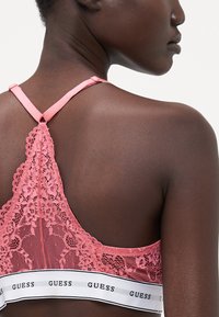 Bralette rosa in pizzo con un delicato motivo floreale, spalline regolabili e un elastico bianco con logo nella parte inferiore.