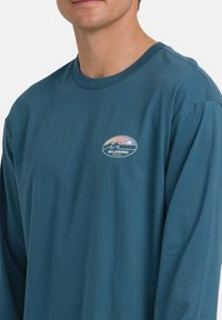 Mężczyzna w turkusowej koszuli z długim rękawem z małym kolorowym logo Billabong przedstawiającym fale i zachód słońca na lewej części klatki piersiowej.