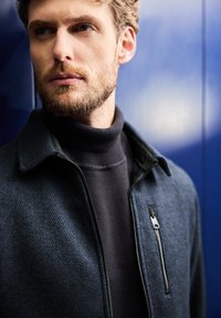 Marinblå texturerad jacka med en dragkedjeförsedd bröstficka, bärs över en mörk polotröja. Bakgrunden visar en mörkblå vägg.