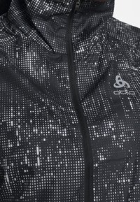 Chaqueta impermeable negra con un patrón pixelado en blanco, que presenta un cuello alto, cremallera frontal completa y un logo en el pecho.