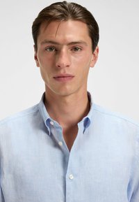 Jasnoniebieska koszula zapinana na guziki z kołnierzykiem, wykonana z teksturowanej tkaniny. Ma widoczne szwy i zaokrąglone mankiety.