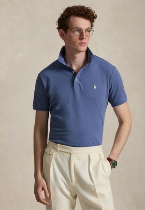 Jaunas vyras su garbanotais plaukais ir akiniais, dėvintis mėlyną polo marškinėlį ir kreminės spalvos plisė kelnes, stovintis su viena ranka kišenėje.