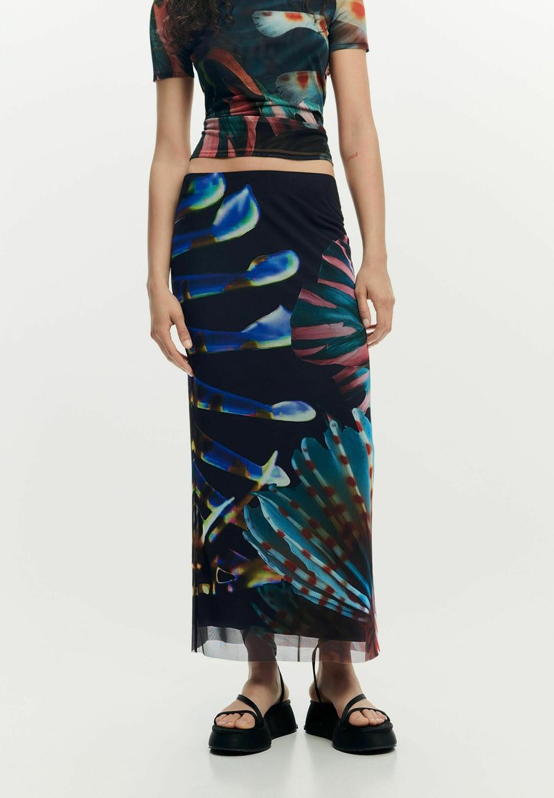 Mujer vistiendo una falda larga y una blusa de manga corta a juego con patrones florales abstractos y coloridos, y sandalias negras de plataforma.