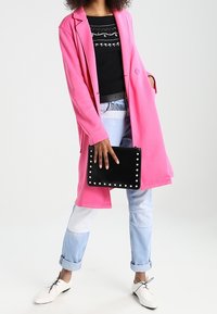Manteau long rose vif, pull noir orné, jean bleu clair avec patchwork, pochette noire avec accents en clous, chaussures blanches.