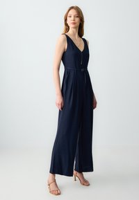 Jumpsuit blu navy con scollatura a V profonda, vita aderente accentuata da una cintura e pantaloni a gamba larga. Tessuto leggero e design pulito.