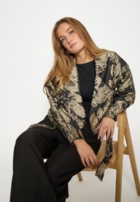 Potlačená bunda v béžovej a čiernej saténovej farbe s voľným strihom, so širokými rukávmi a kontrastným čiernym vrchom. Voľné čierne nohavice.