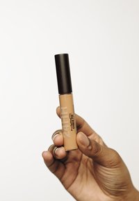 24-Stunden Studio Fix Concealer in einer klaren Tube mit einem mattschwarzen Deckel, der einen beigen Farbton zeigt und silberne Schriftzüge an der Seite hat. Eine Hand hält das Produkt.