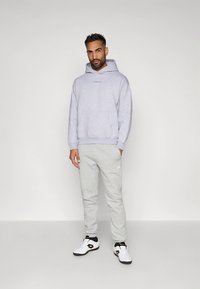 Felpa con cappuccio grigio chiaro con tasca frontale e logo, abbinata a pantaloni della tuta grigio chiaro coordinati, completata da sneakers bianche con dettagli neri.