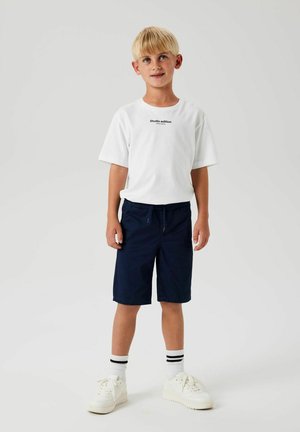 T-shirt blanche avec l'impression "édition studio", shorts navy, baskets blanches et chaussettes rayées, se tenant devant un fond uni.